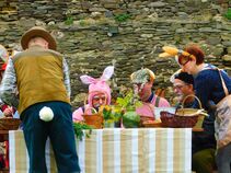 Gruppe von Erwachsenen in Hasenkostümen und mit Hasenohren sitzt an einem Tisch mit Körben und Frühlingsdeko. Eine Frau reicht etwas über den Tisch, während ein Mann mit Hasenschwanz den Tisch bedient.