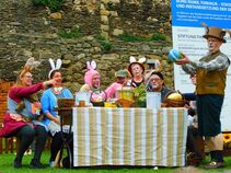 Mann in Hasenkostüm hält ein großes buntes Osterei hoch. Am dekorierten Ostertisch sitzen mehrere verkleidete Erwachsene mit Hasenohren und Gesichtsbemalung.