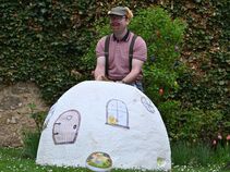 Mann mit Hasenohren und bemaltem Gesicht steht hinter einem bemalten weißen Iglu-artigen Aufbau im Garten. Im Hintergrund ein Strauch mit bunten Ostereiern.