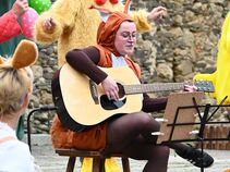 Desiree Meinhardt im Eichhörnchenkostüm spielt Gitarre auf einem Hocker. Um sie herum stehen Darsteller in bunten Tierkostümen.