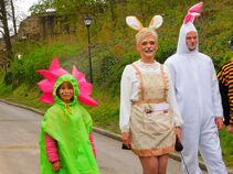 Drei Personen in bunten Kostümen beim Osterspaziergang: ein Kind als Blume, eine Frau als Häsin und ein Mann im Hasenkostüm, im Hintergrund eine Burgmauer.