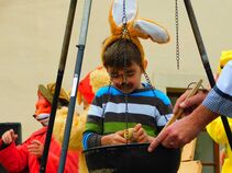 Junge mit Hasenohren und bemaltem Gesicht steht an einem Hängekessel und bemalt ein Ei. Ein Erwachsener zeigt ihm etwas, während im Hintergrund weitere Kinder in Tierkostümen zusehen.