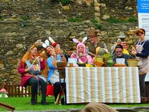Fröhliche Szene an einem gedeckten Tisch: Mehrere Erwachsene in bunten Hasenkostümen sitzen vor einer Steinmauer, lachen, basteln und bemalen Eier. Dekorierte Körbe und Frühlingsblumen stehen auf dem Tisch.