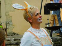 Frau mit kurzen blonden Haaren, Hasenohren und bemaltem Gesicht steht lächelnd vor einem Hängekessel mit Holzfeuer.