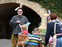 Mann mit Hasenohren steht vor dem Burgtor und schaut auf Kinder in Hasenkostümen mit Rucksäcken. Eine Frau mit Schürze ist ebenfalls zu sehen.