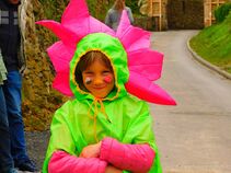 Ein Kind in einem leuchtend grünen Blumen-Kostüm mit pinken Blütenblättern und bemaltem Gesicht lächelt in die Kamera.