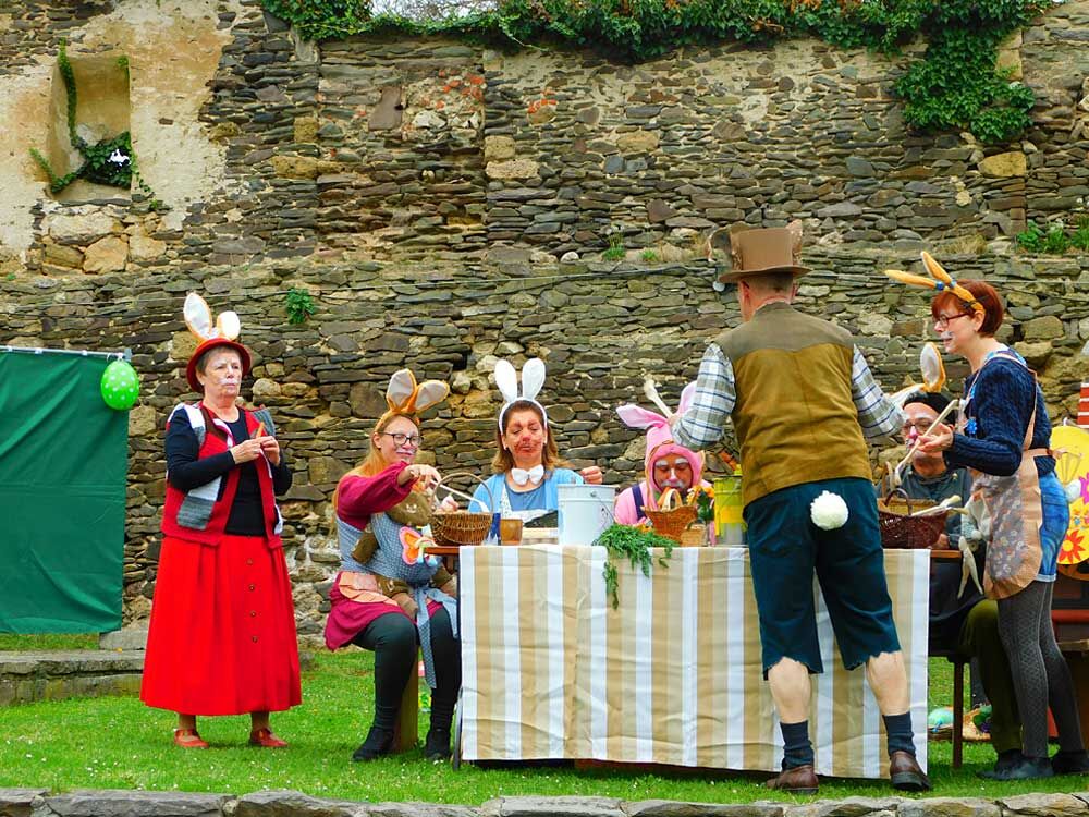 Große Gruppe von als Hasen verkleideten Personen sitzt und steht an einem geschmückten Tisch im Burghof der Burg Ranis, alle inmitten einer fröhlichen Spielszenen.