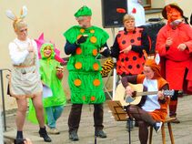 Gruppe verkleideter Darsteller tanzt und musiziert auf der Bühne. Ein Mädchen im Blütenkostüm steht zwischen Erwachsenen in Hasen-, Marienkäfer- und Käferkostümen.