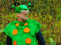 Ein Mann im grünen Raupenkostüm mit orangefarbenen Punkten, Fühlern und übergroßer Brille kaut auf einem Blatt, im Hintergrund dichte Sträucher.