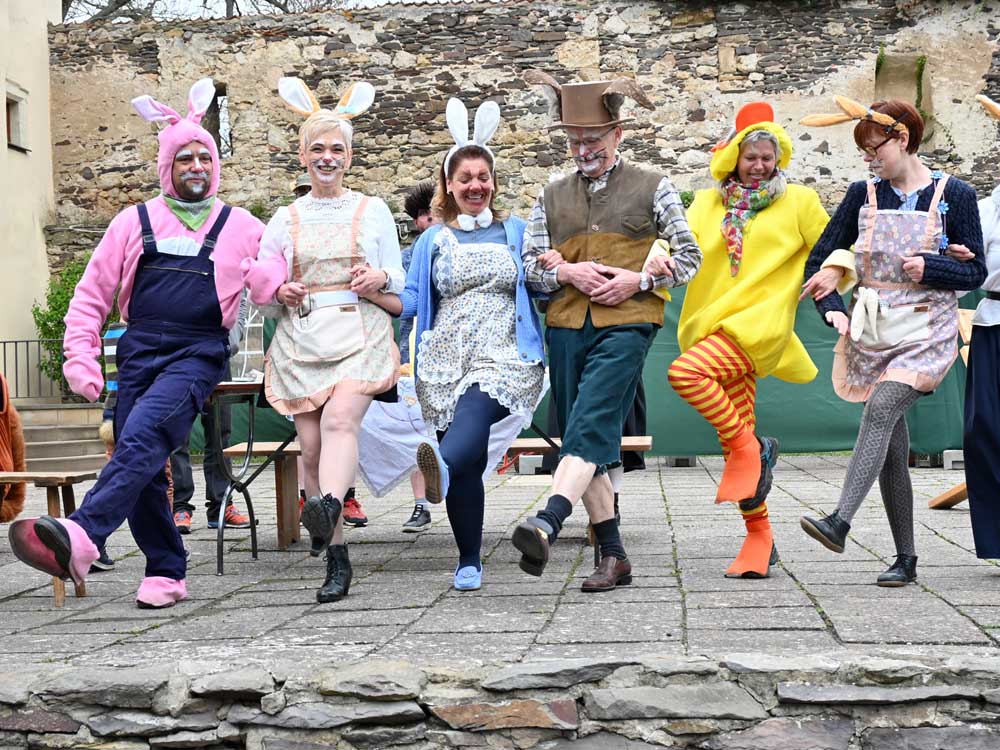 Verkleidete Darsteller tanzen in einer Reihe mit eingehakten Armen. Sie tragen bunte Osterkostüme, darunter Hasen, Huhn und Koch. Im Hintergrund eine alte Burgmauer.